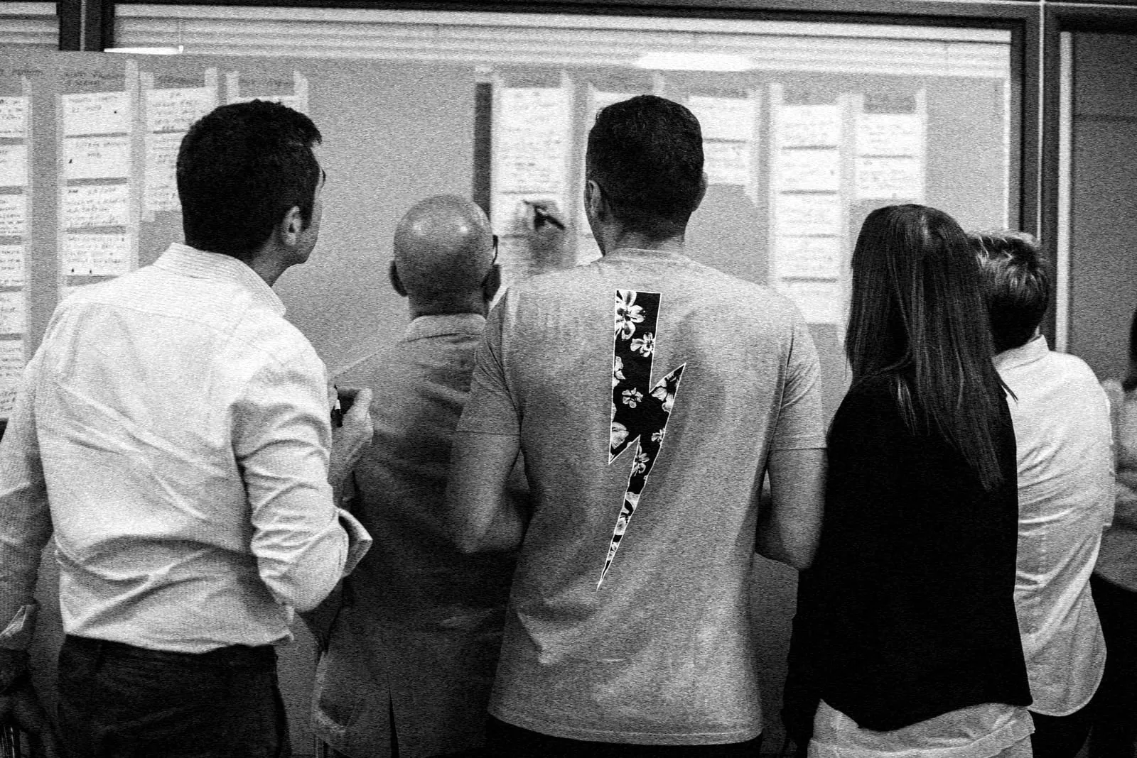 Ein Team steht vor einer Wand mit Notizzetteln; ein Mann schreibt etwas an, die anderen schauen zu.